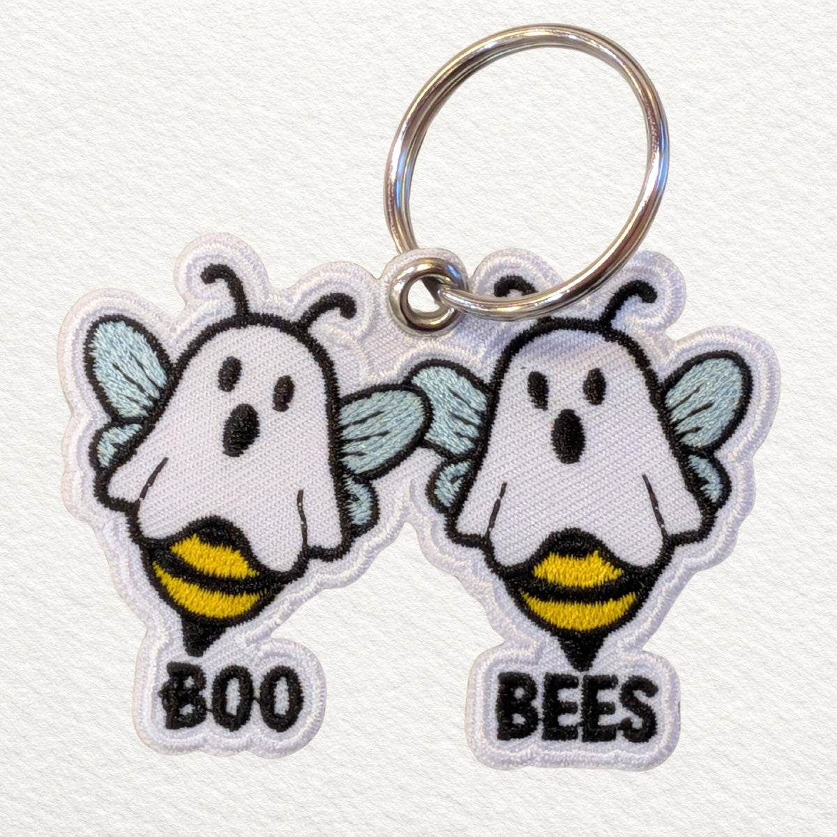 Two embroidered keychain charms shaped like a ghost bees, attached to a silver metal key ring on a white textured background. One ghost bee says 'BOO' and the other says 'BEES'.