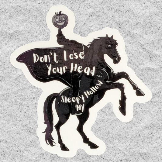 A die-cut vinyl sticker featuring a black silhouette of The Headless Horseman on a rearing horse, holding a carved pumpkin. The words 'Don't Lose Your Head' and 'Sleepy Hollow NY' are printed in white script across the design, set against a textured gray background.