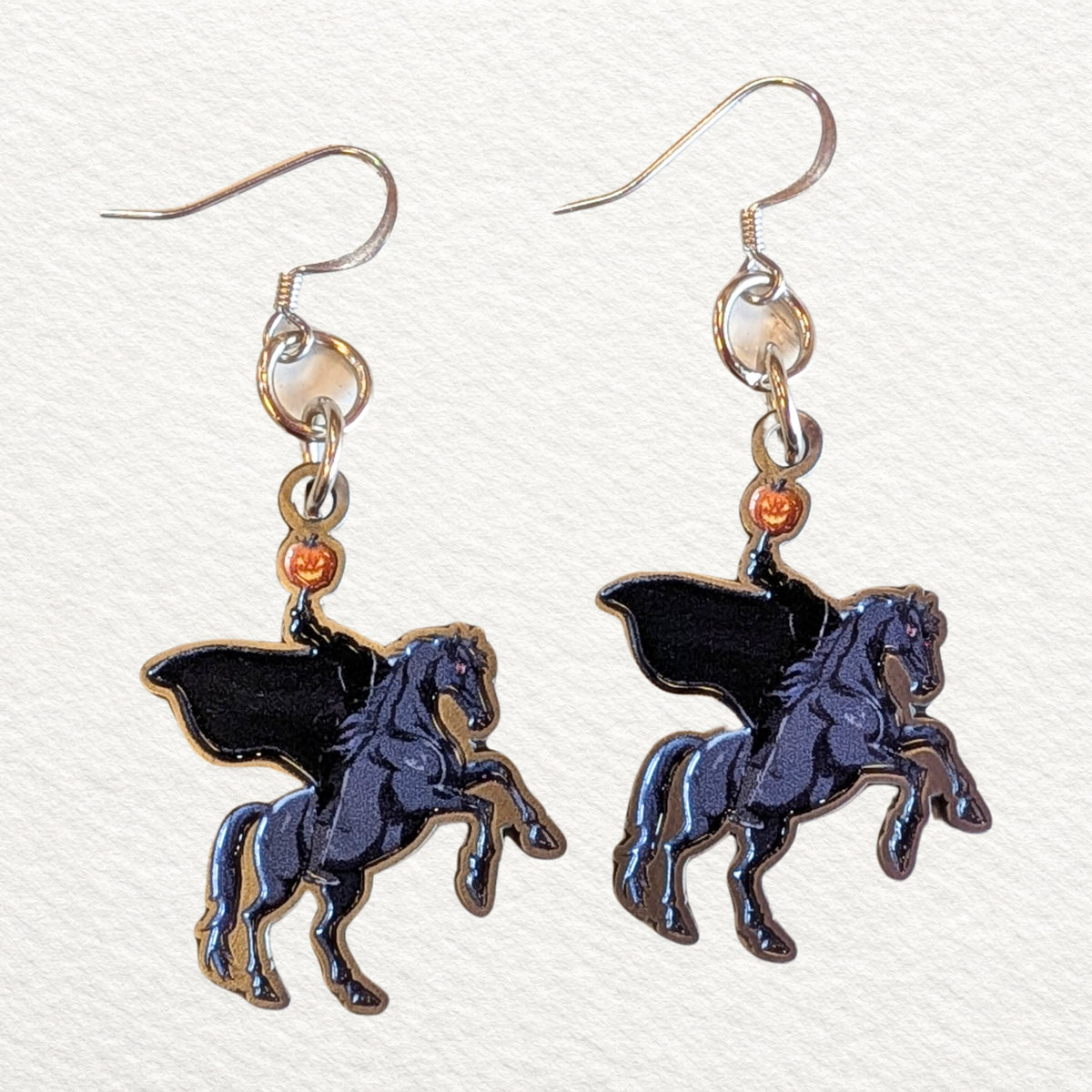 A pair of dangling earrings featuring a glossy headless horseman, suspended from stainless steel fishhook wires, set against a textured off-white background.