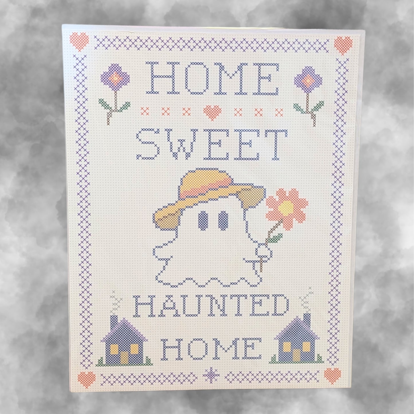 A cross-stitch style decorative print reading “HOME SWEET HAUNTED HOME,” featuring a ghost in a yellow hat holding a flower, with small houses and flowers around the border on a light background.