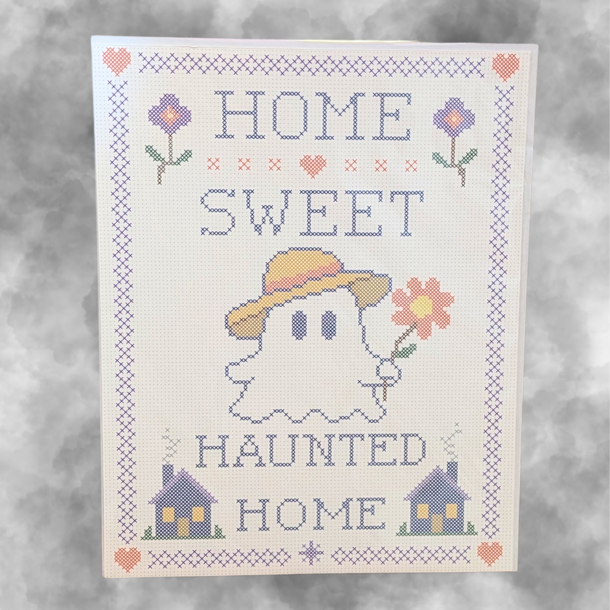 A cross-stitch style decorative print reading “HOME SWEET HAUNTED HOME,” featuring a ghost in a yellow hat holding a flower, with small houses and flowers around the border on a light background.