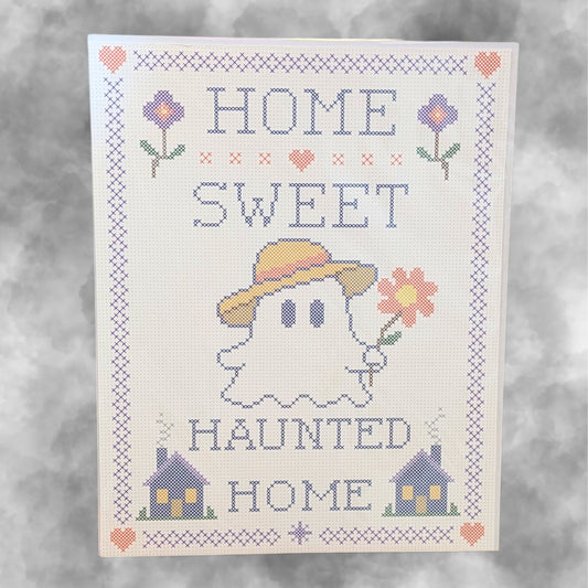 A cross-stitch style decorative print reading “HOME SWEET HAUNTED HOME,” featuring a ghost in a yellow hat holding a flower, with small houses and flowers around the border on a light background.
