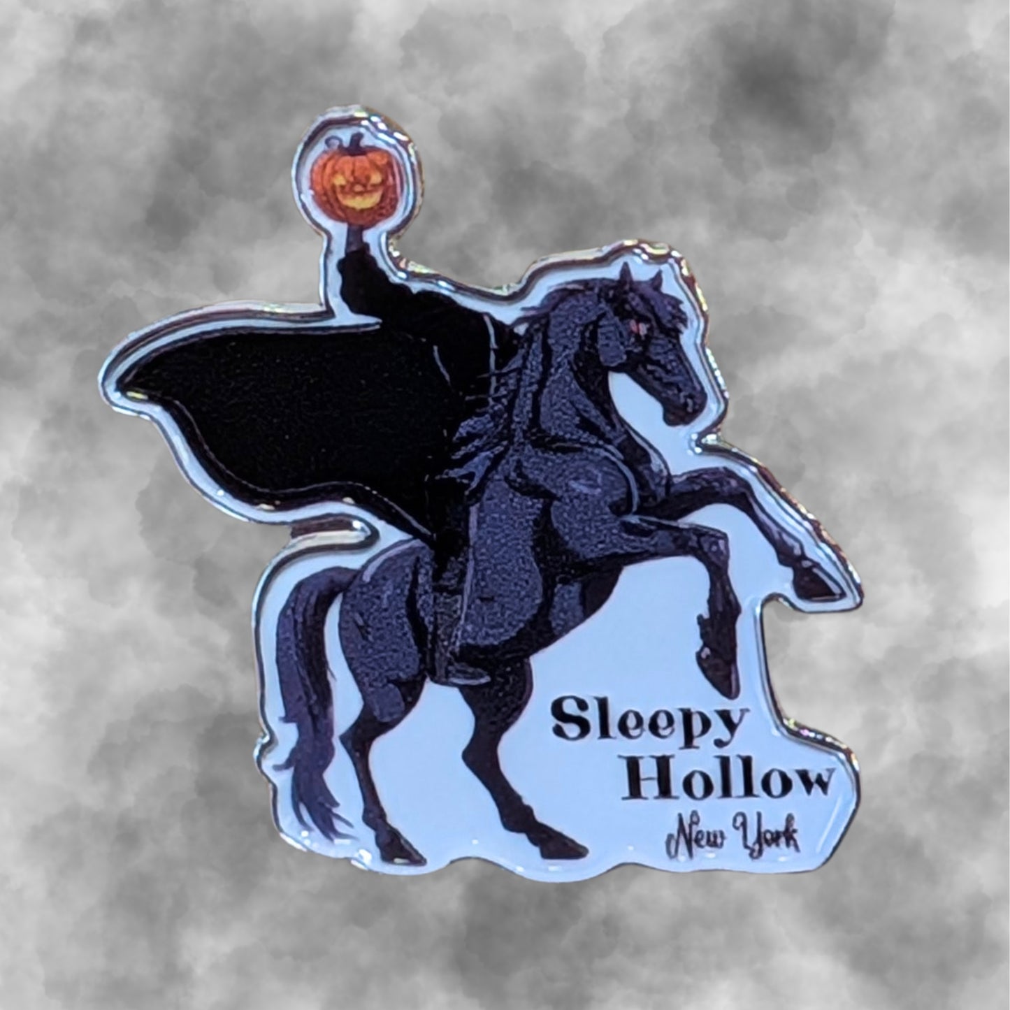 A die-cut enamel pin featuring a black silhouette of the Headless Horseman on a rearing black horse, holding a glowing orange jack o-lantern. The pin has a smokey grey background with the text 'Sleepy Hollow New York' in black.