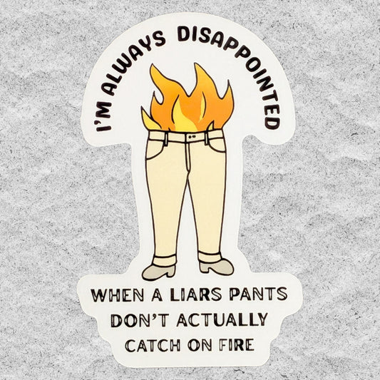 A die-cut sticker featuring a cartoon of beige pants on a white background with yellow and orange flames rising from the waist area. The sticker has black text that reads 'I'M ALWAYS DISAPPOINTED WHEN A LIARS PANTS DON'T ACTUALLY CATCH ON FIRE' and is shown on a textured gray background.