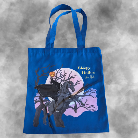 A navy blue canvas tote bag with a Halloween-themed graphic print featuring a dark knight on a horse against a pink moon and bare trees, with the text 'Sleepy Hollow New York'.