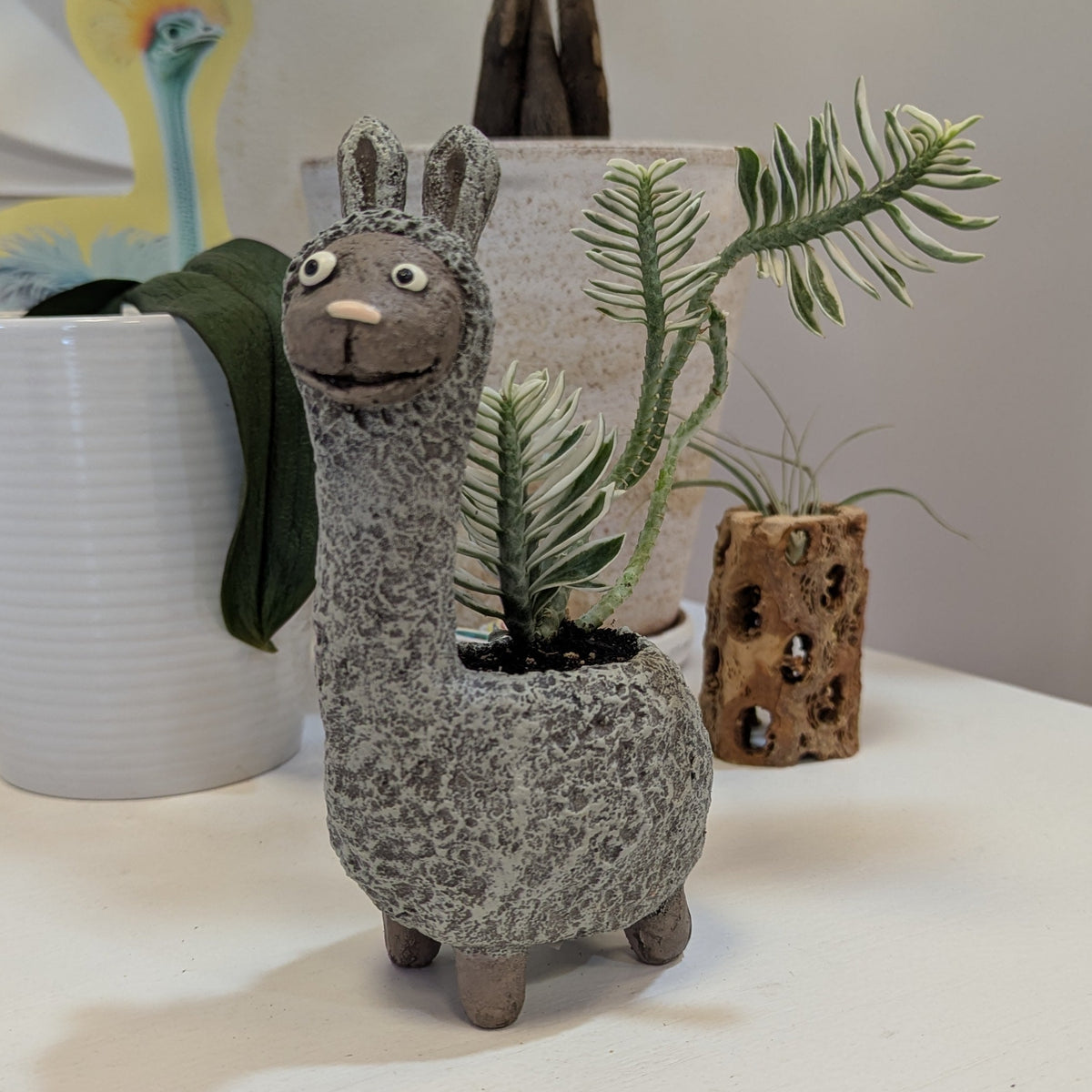 Ceramic llama-shaped planter with a plant inside, placed on a white surface with decorative items in the background.