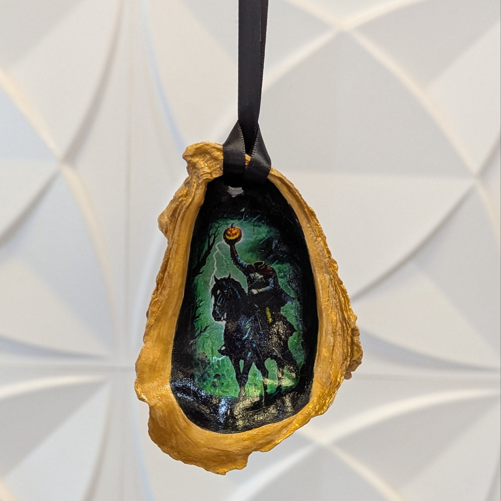 Hand crafted ornament on an oyster shell with an image of the Headless Horseman on a green background and gold edges hanging from black ribbon.