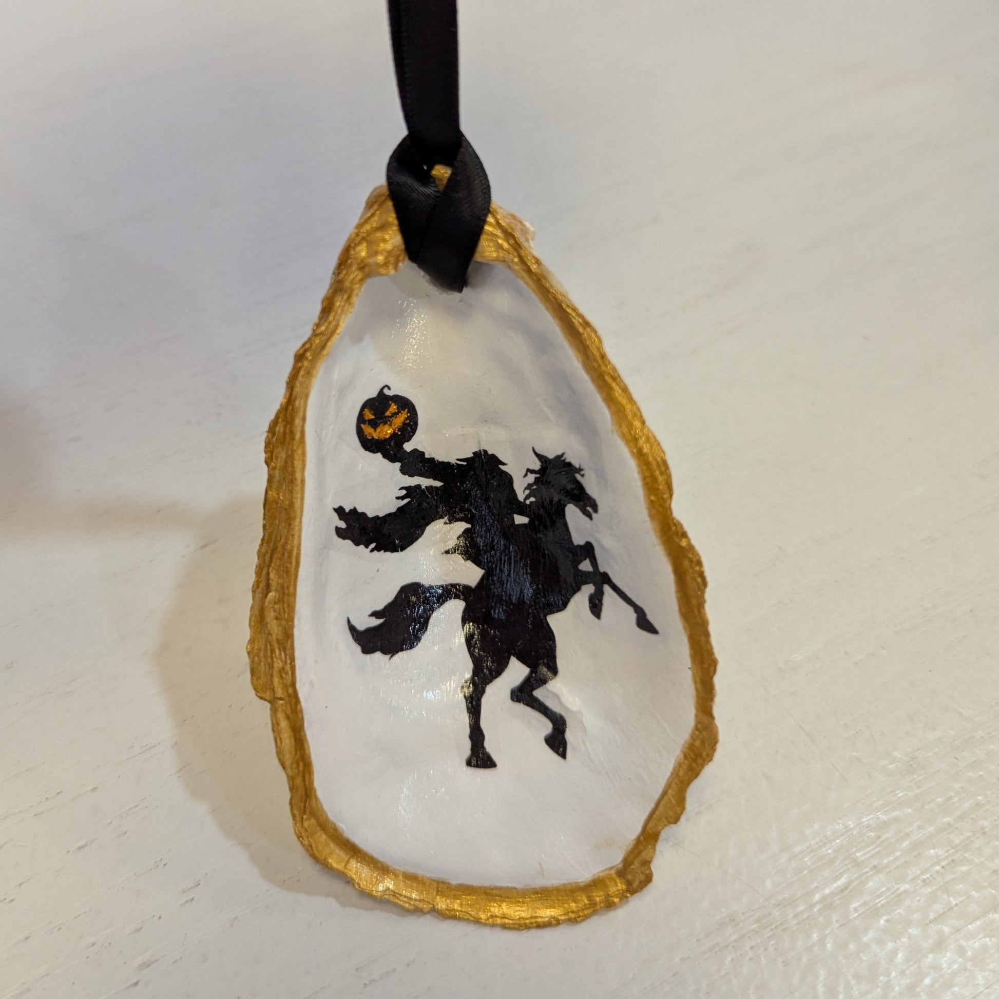 Hand crafted ornament on an oyster shell with an image of the Headless Horseman on a white background and gold edges hanging from black ribbon.