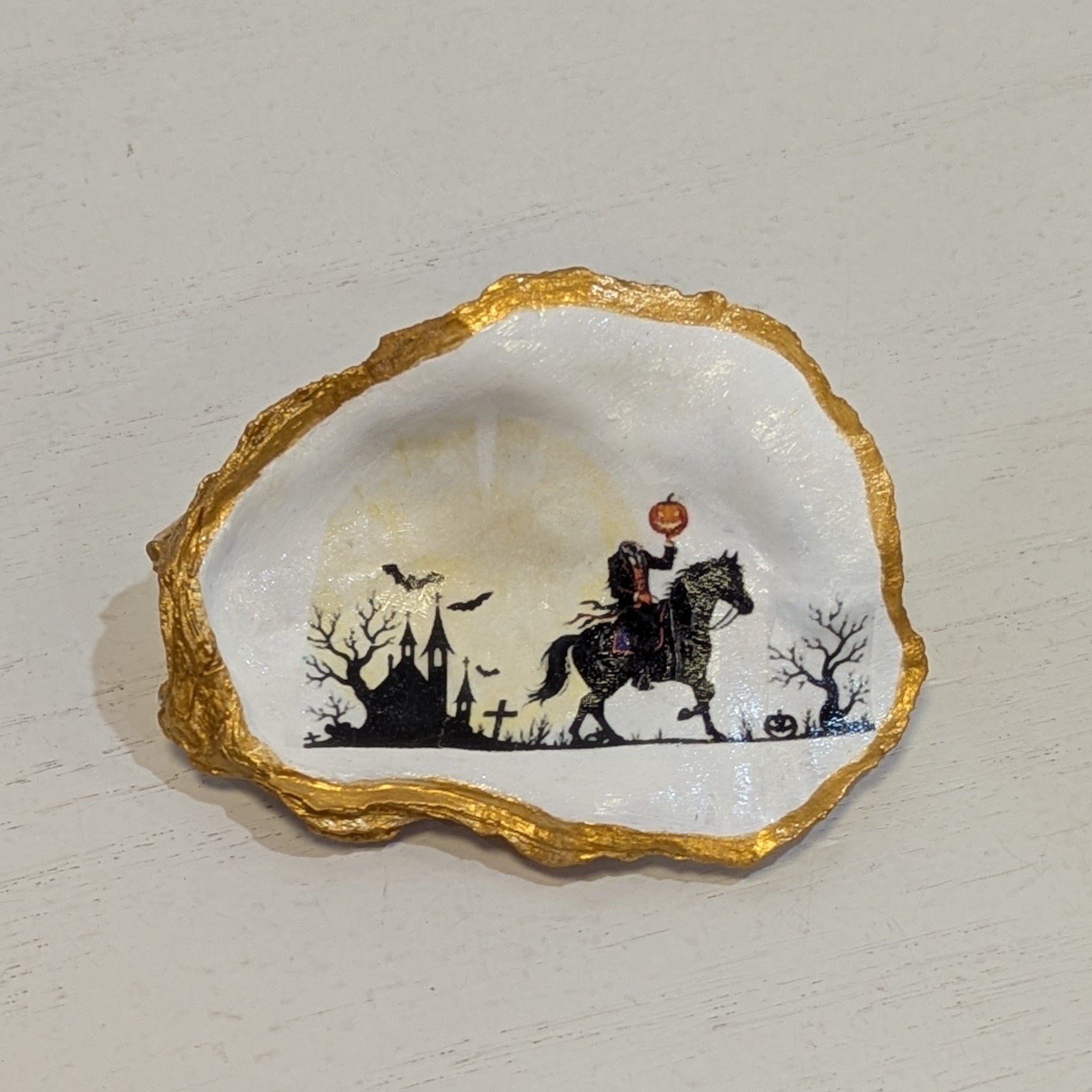 Hand crafted ornament on an oyster shell with an image of the Headless Horseman on a white background and gold edges sitting on a white surface.