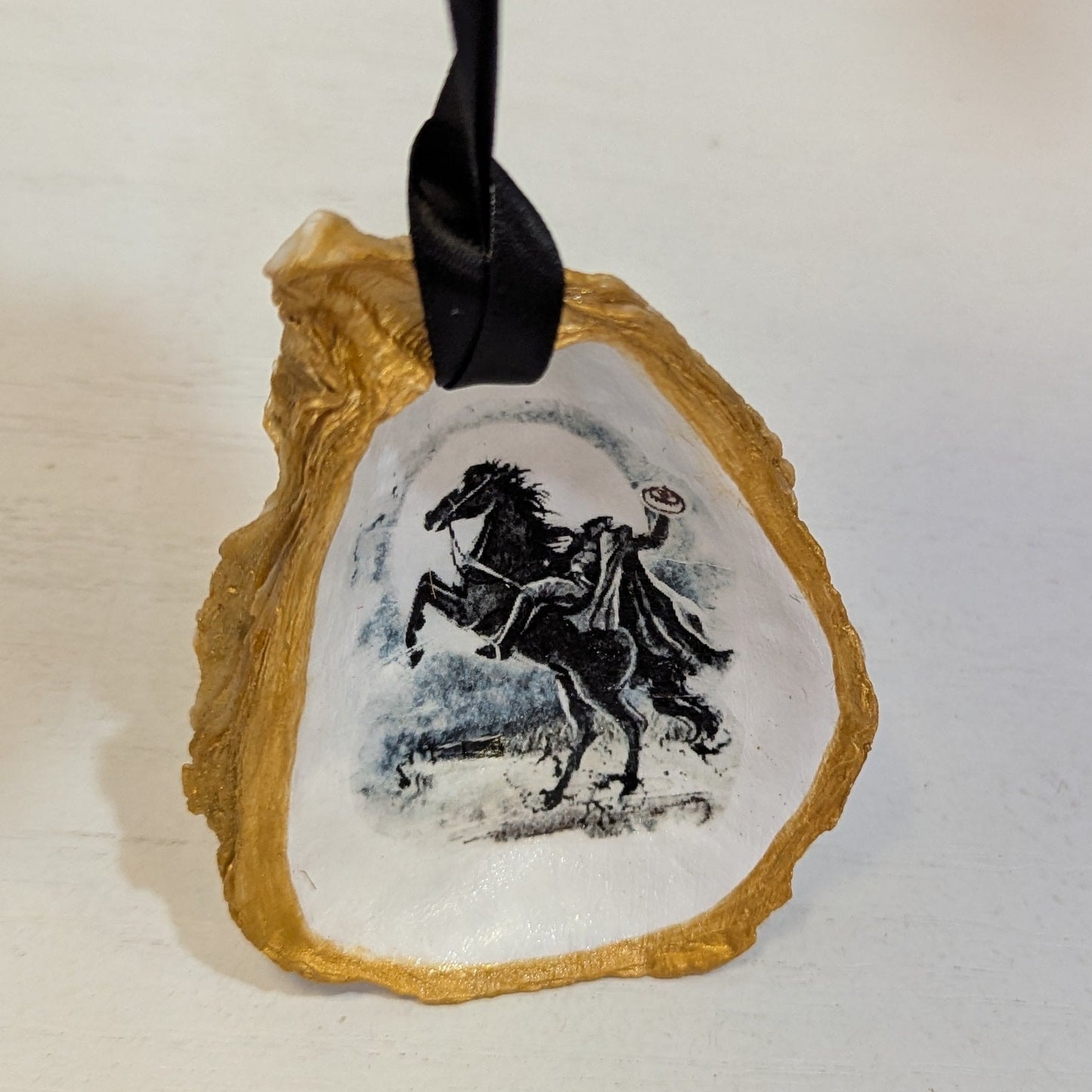 Hand crafted ornament on an oyster shell with an image of the Headless Horseman on a white background and gold edges hanging from black ribbon.