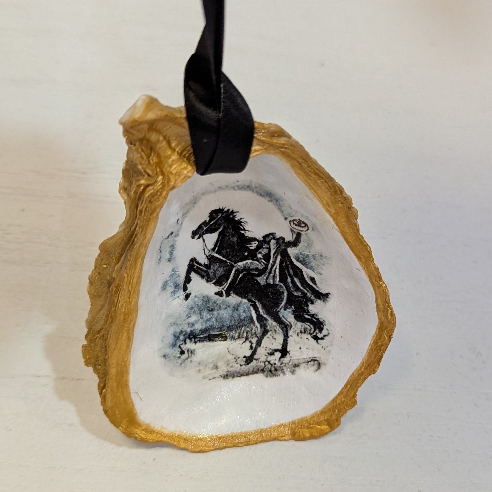 Hand crafted ornament on an oyster shell with an image of the Headless Horseman on a white background and gold edges hanging from black ribbon.
