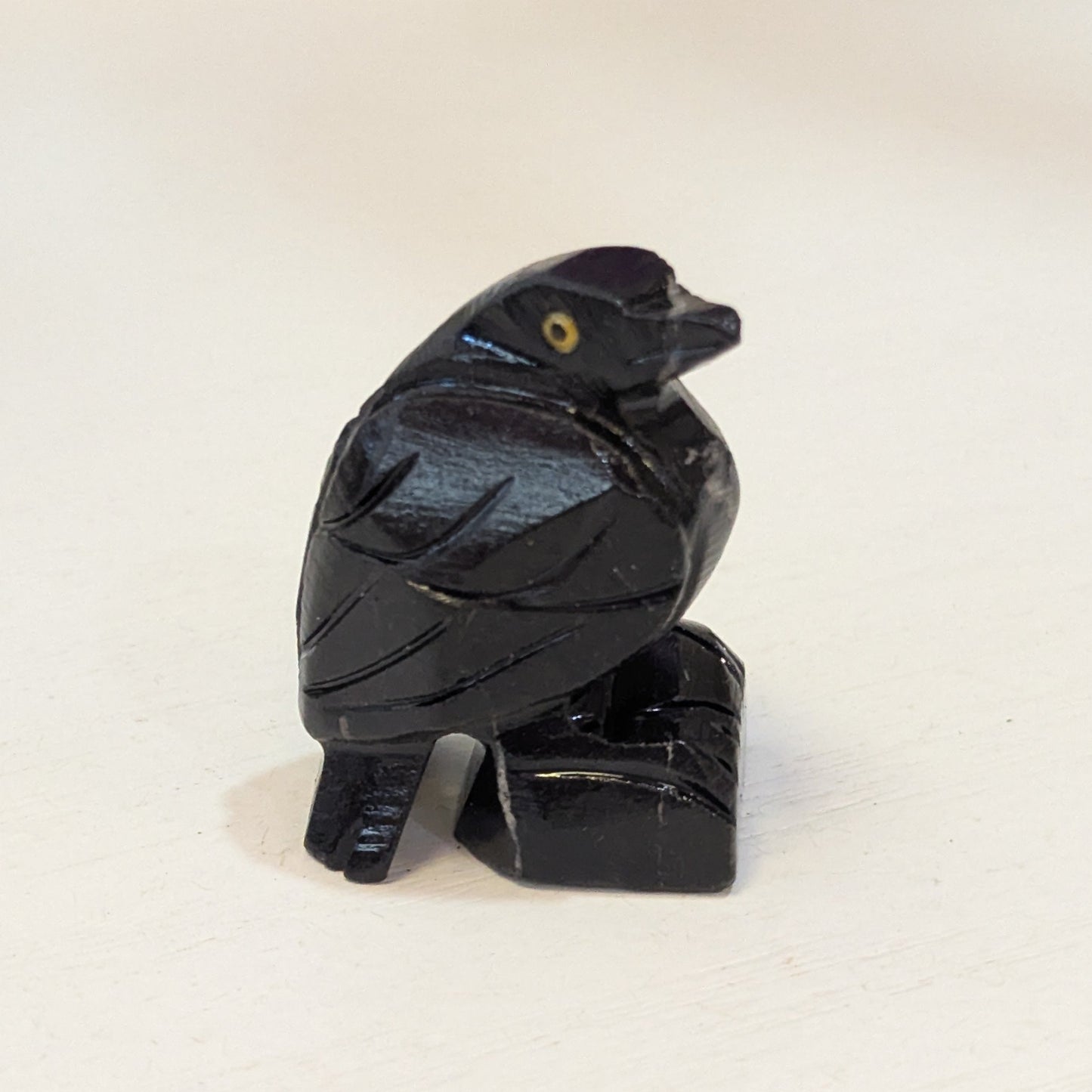 A small carved black Raven figurine sitting on a small black base, photographed on a light cream surface.