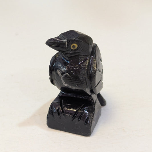 A small carved black Raven figurine, standing on a rectangular base. The bird has a glossy black finish with a small yellow eye detail, photographed on a plain off-white surface.