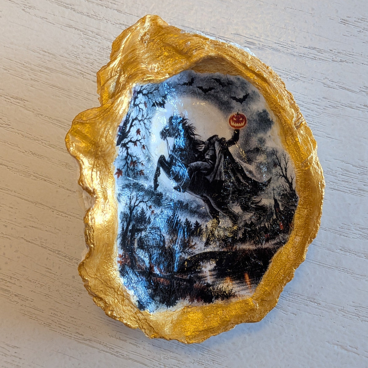 A decorative Oyster Shell with a textured gold border and a hand-painted central scene of The Headless Horseman on a snowy landscape, displayed on a light wood surface.