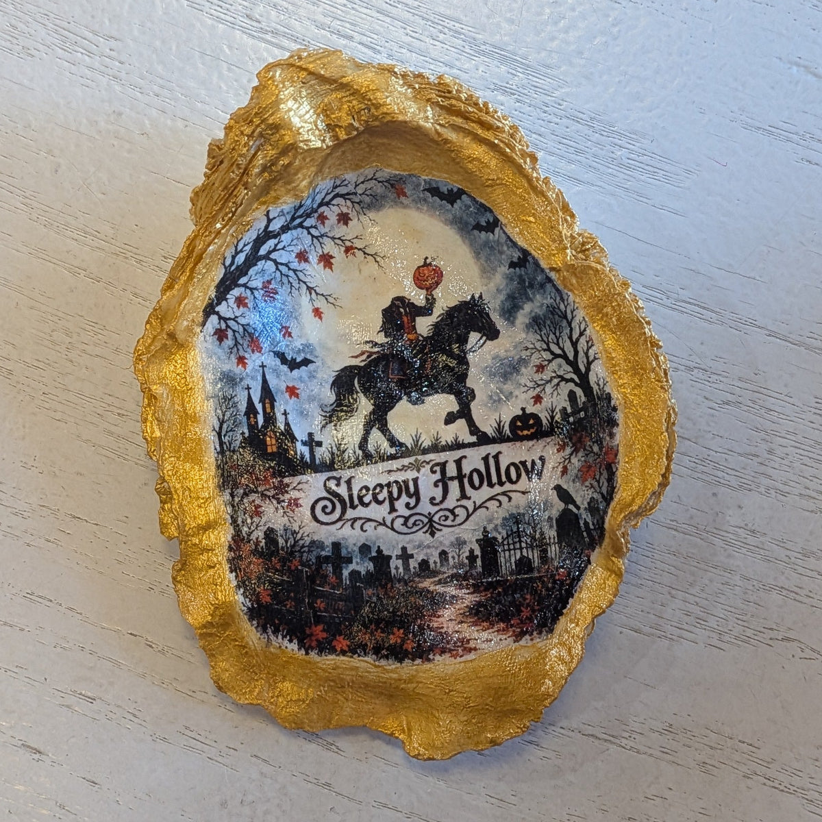 A small decorative Oyster Shell with a gold-toned irregular border and a printed scenic insert reading 'Sleepy Hollow,' placed on a light wood tabletop.
