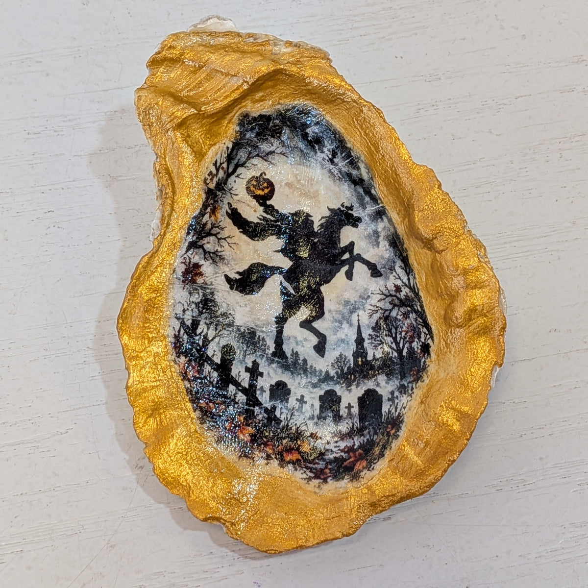 A small decorative oyster shell piece with a gold-toned rim and a central painted scene of a headless horseman on a horse against a snowy night sky, placed on a light off-white surface.