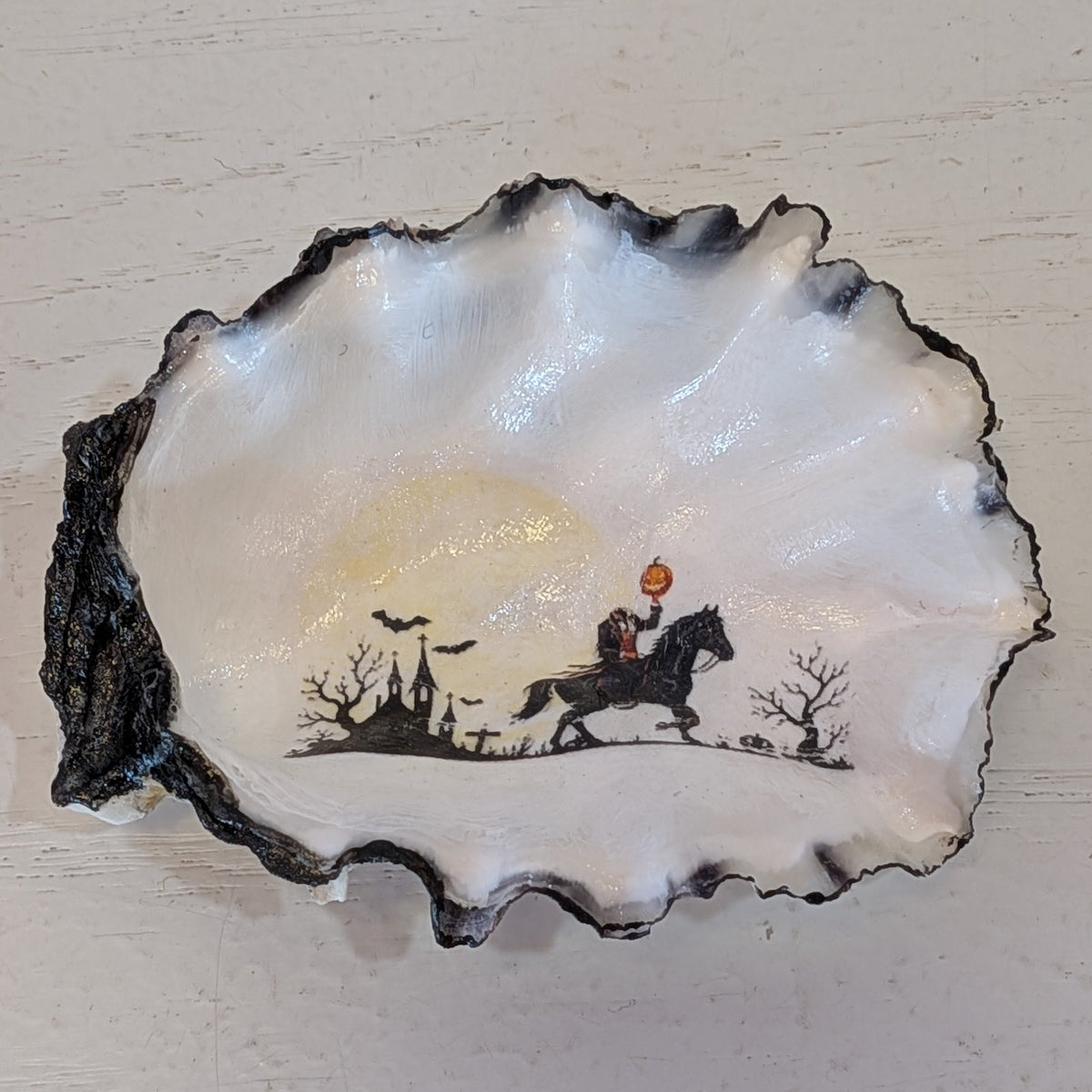 A sitting decoration shaped like an open oyster shell, featuring a black and white painted scene of a headless horseman on a horse under a glowing yellow moon, set against a light textured background.