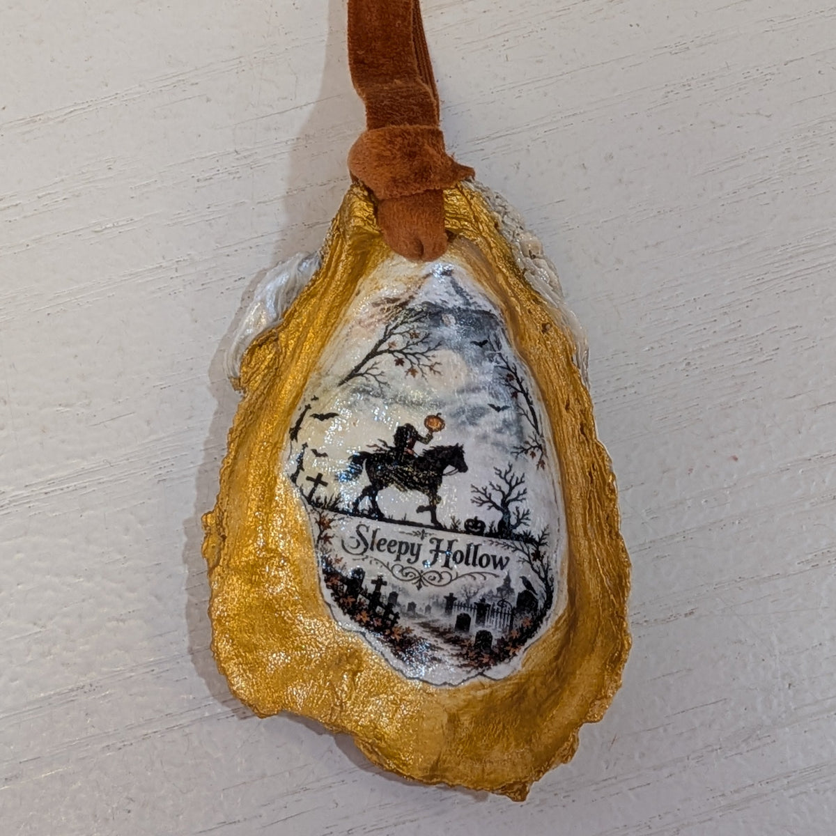 A decorative holiday ornament shaped like a natural oyster shell with a brown felt hanging loop. The shell is gold on the sides and white in the center, featuring a central printed illustration of a snowy winter landscape with a horse rider and the text 'Sleepy Hollow'
