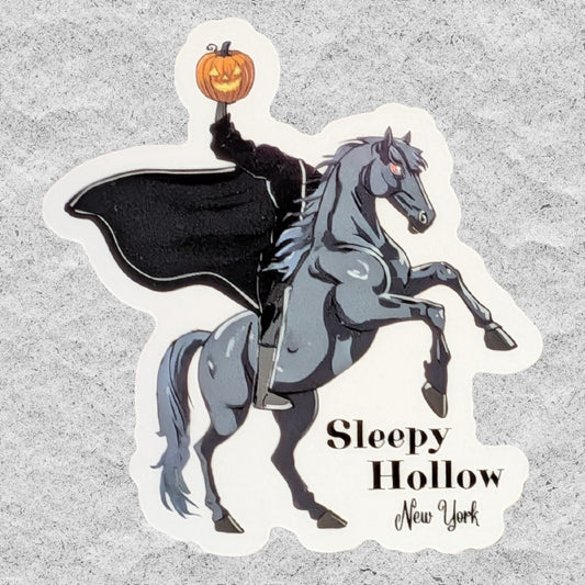 A die-cut sticker featuring an illustration of a gray horse rearing up while being ridden by The Headless Horseman in a black cloak who is holding up a carved orange pumpkin. The text 'Sleepy Hollow New York' is printed on the sticker, which is displayed on a textured gray background.