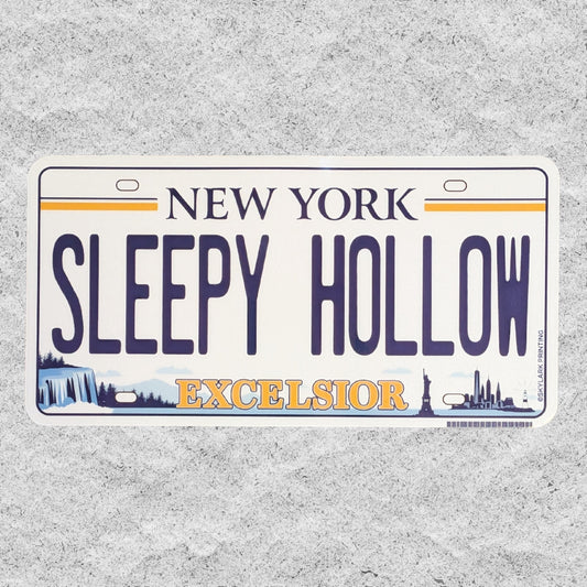 A die-cut sticker of a license plate styled like a New York plate, reading 'SLEEPY HOLLOW' with 'EXCELSIOR' below, photographed on a textured light gray background.
