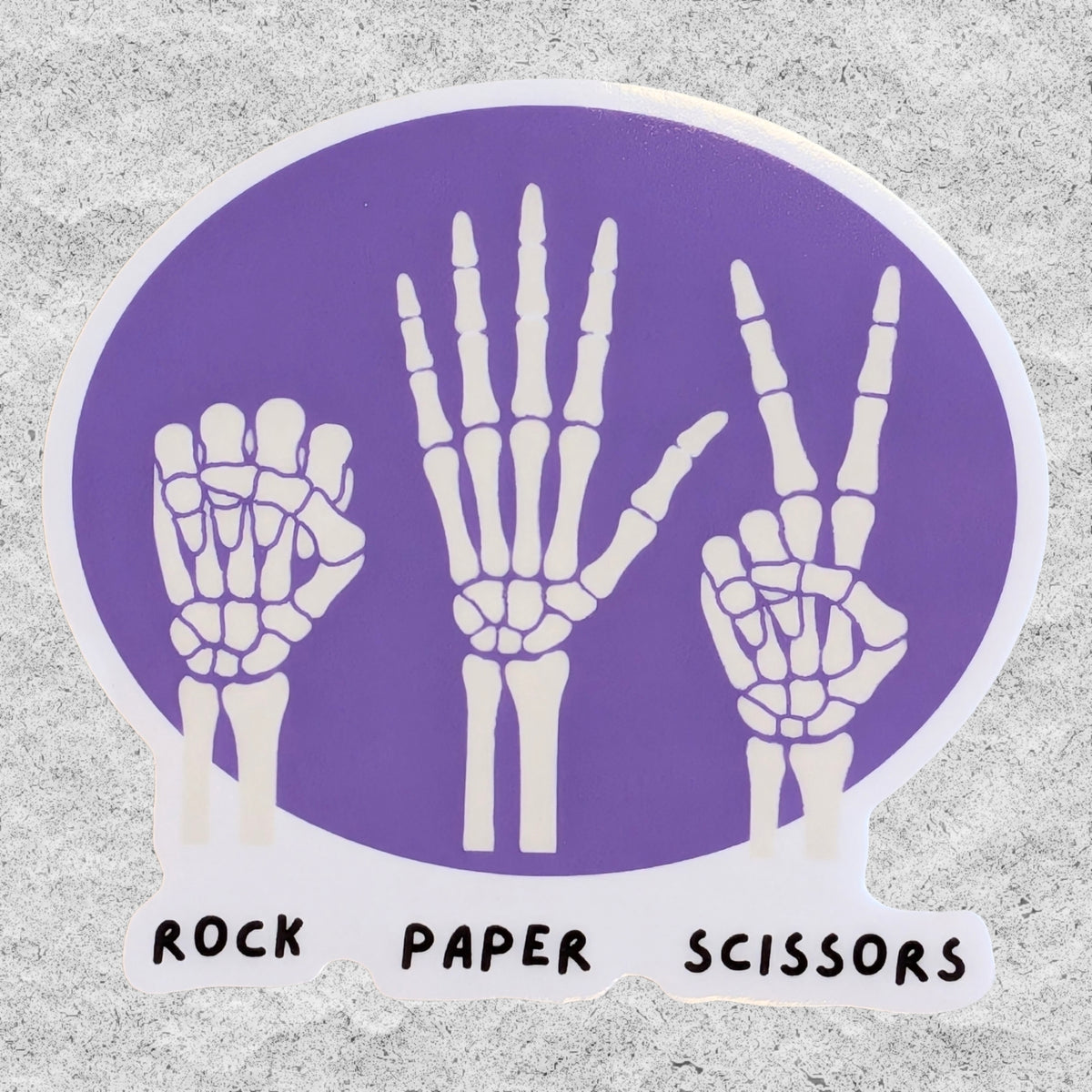 A sticker with three skeleton hands doing rock, paper, scissors on a purple background.