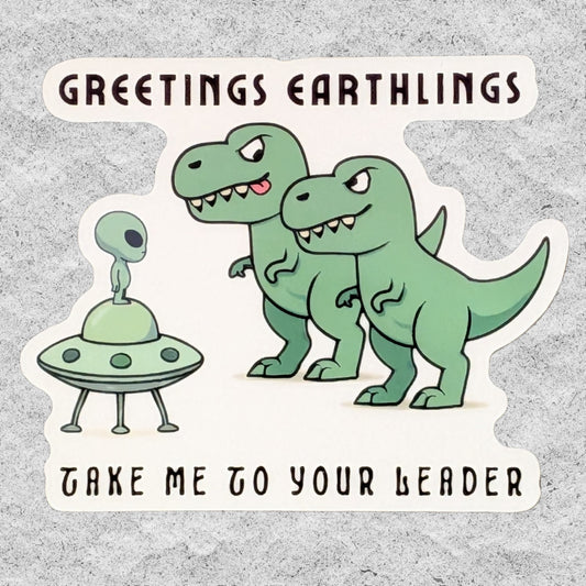 A die-cut sticker featuring a cartoon scene with two hungry green T-Rex dinosaurs and a small green alien in a UFO, with the text 'GREETINGS EARTHLINGS' at the top and 'TAKE ME TO YOUR LEADER' at the bottom, shown on a textured light gray background.