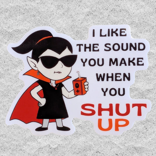 A die-cut vinyl sticker featuring a cartoon vampire character in a black dress and cape holding an orange juice box, next to the text 'I LIKE THE SOUND YOU MAKE WHEN YOU SHUT UP' in black and orange lettering, shown on a textured gray background.