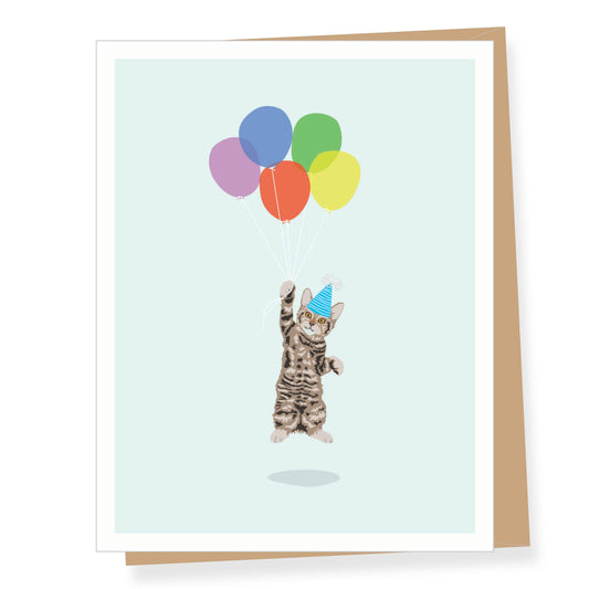 A greeting card with a white border showing an illustration of a small cat wearing a party hat and reaching up to colorful balloons, with a tan envelope behind it on a white background.