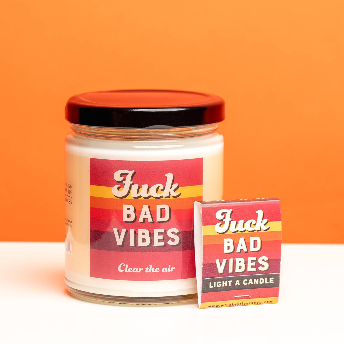 A jar candle labeled 'Juck Bad Vibes Clear the air' with a red lid, shown next to its matching rectangular safety matches on a white surface against an orange background.