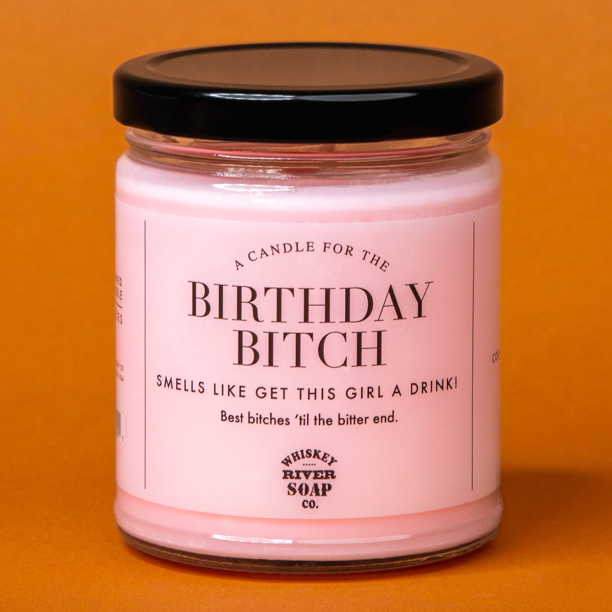 A pink candle in a clear glass jar with a black lid, featuring a white label with black text that reads 'A CANDLE FOR THE BIRTHDAY BITCH' and 'SMELLS LIKE GET THIS GIRL A DRINKI'. The label also shows the brand 'Whiskey River Soap Co.