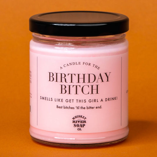 A pink candle in a clear glass jar with a black lid, featuring a white label with black text that reads 'A CANDLE FOR THE BIRTHDAY BITCH' and 'SMELLS LIKE GET THIS GIRL A DRINKI'. The label also shows the brand 'Whiskey River Soap Co.
