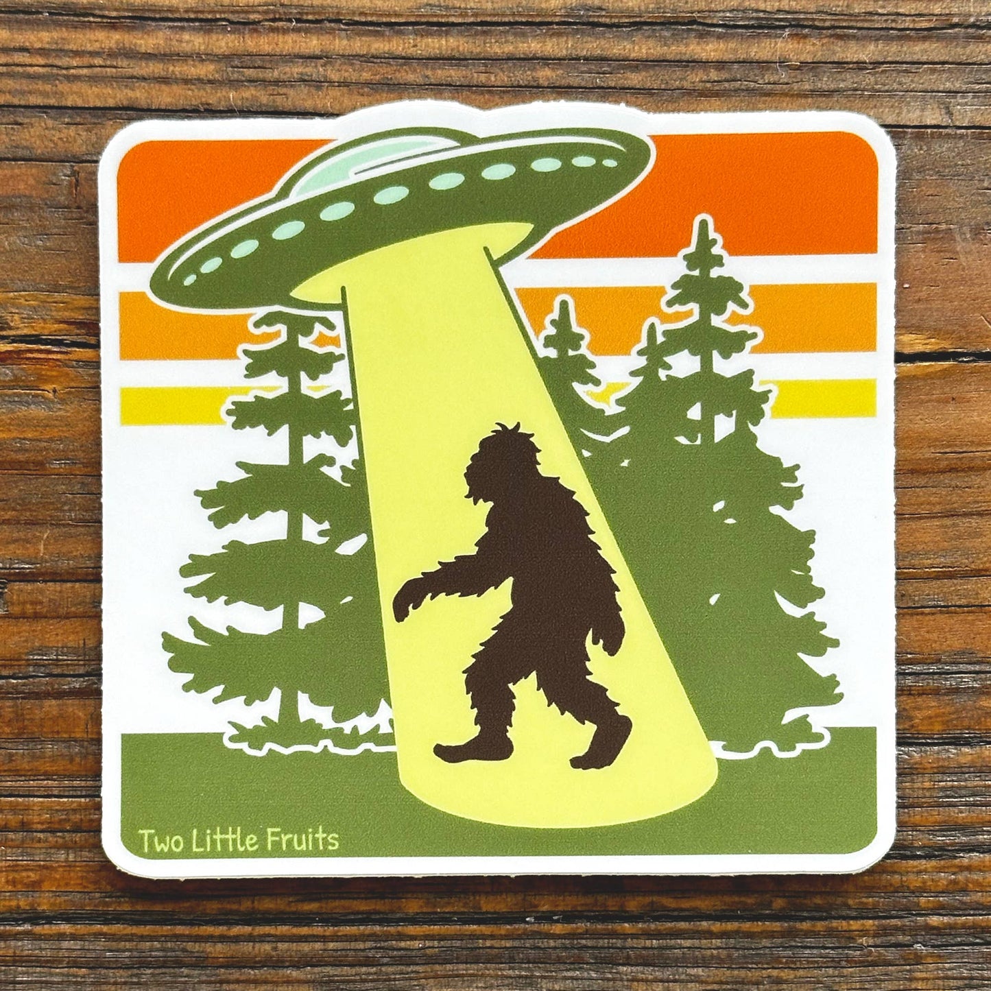 Sticker with a UFO over a silhouette of a bigfoot between trees on a wooden surface.