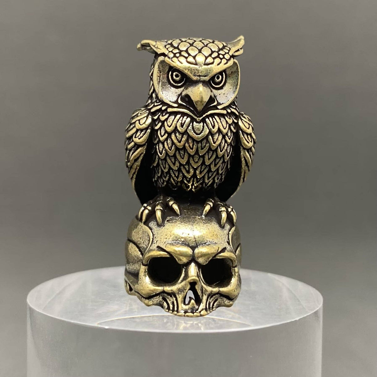 An ornate brass owl figurine perched on a skull, displayed on a clear cylindrical pedestal against a neutral gray background.