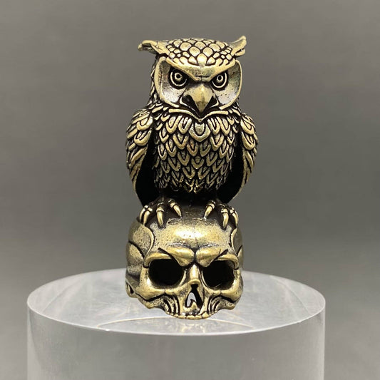 An ornate brass owl figurine perched on a skull, displayed on a clear cylindrical pedestal against a neutral gray background.