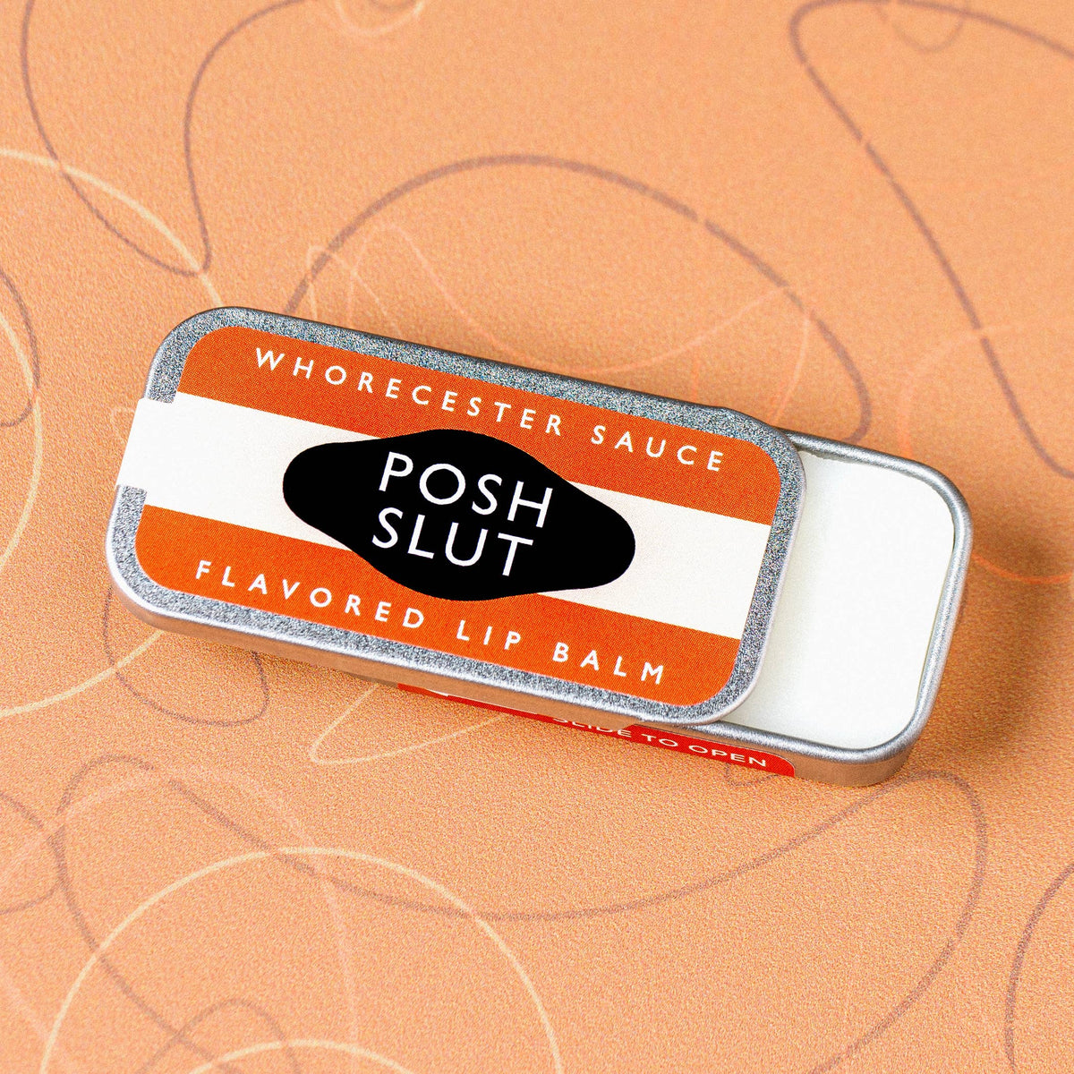 Lip balm tin labeled 'Posh Slut' on an orange background