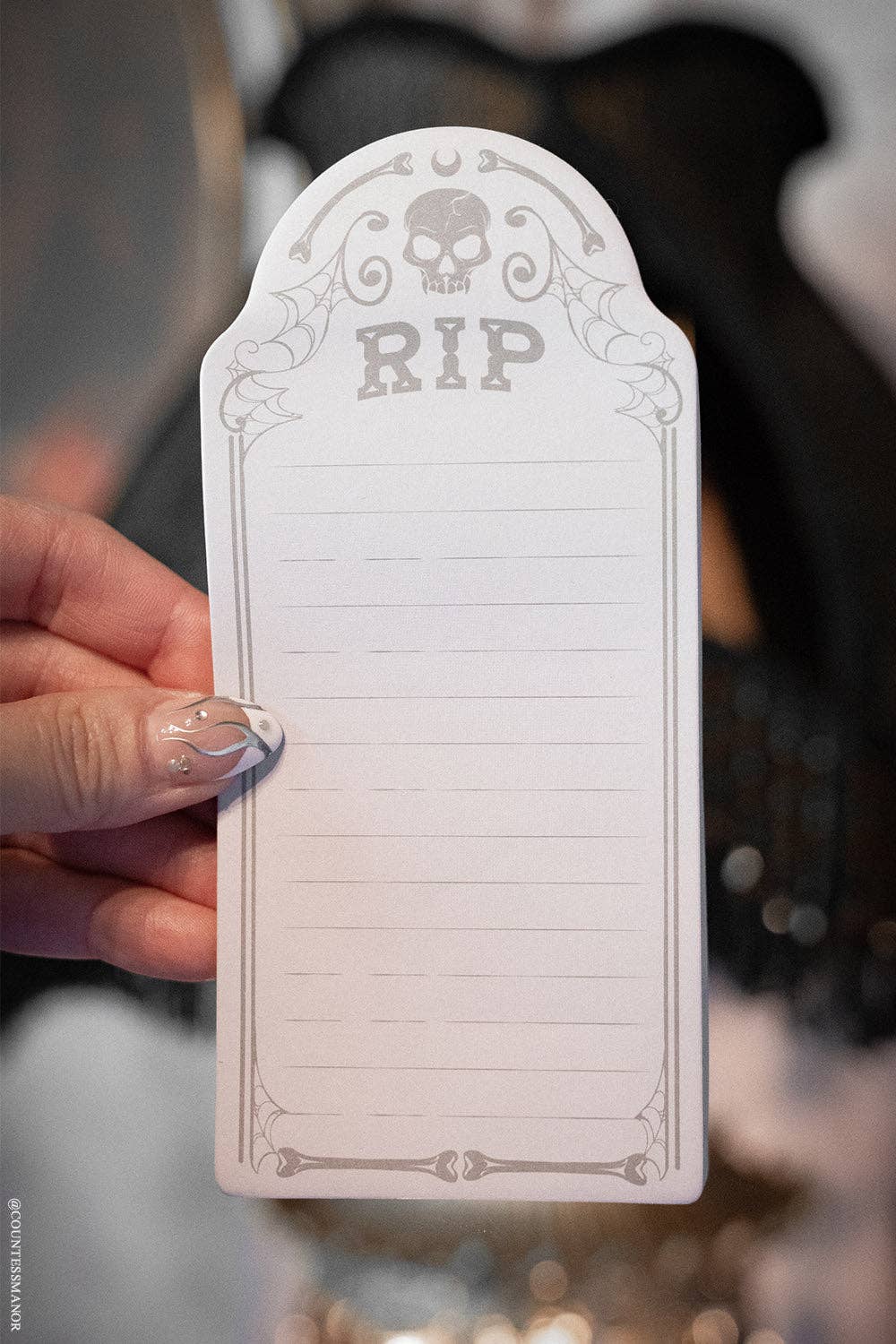 A hand with glittery fingernails holds a white arched notepad featuring a gray 'RIP' skull and spider design on a blurred dark background.