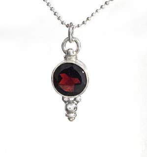 A silver pendant necklace featuring a round dark red gemstone in a textured silver bezel setting, suspended from a silver ball chain against a white background.