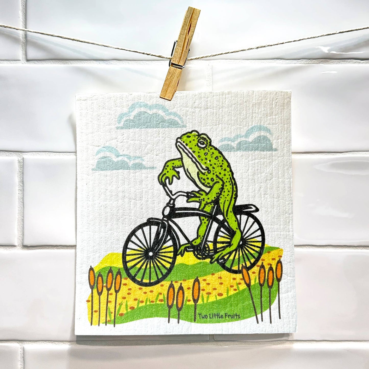 Frogs on a bicycle with a scenic background on a Swedish dish cloth, hanging on a line.