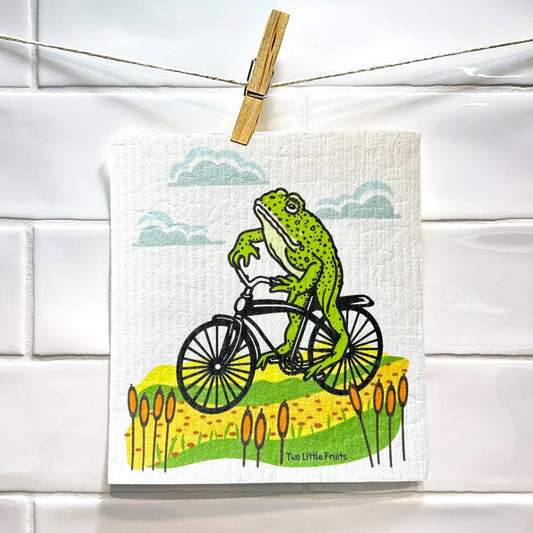 Frogs on a bicycle with a scenic background on a Swedish dish cloth, hanging on a line.