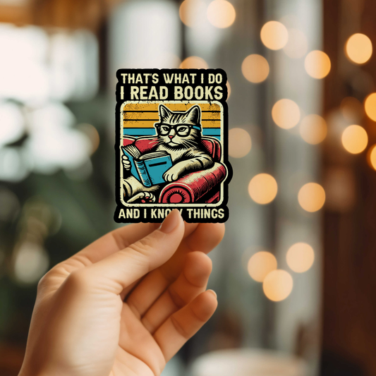 Sticker with a cat reading a book and text 'That's what I do, I read books and I know things' held by a hand against a blurred background.