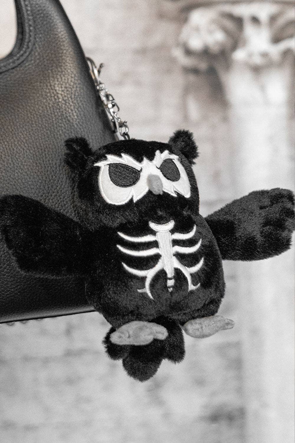 A black plush owl keychain with white skeleton embroidery, hanging from a black leather bag strap against a blurred gray stone background.