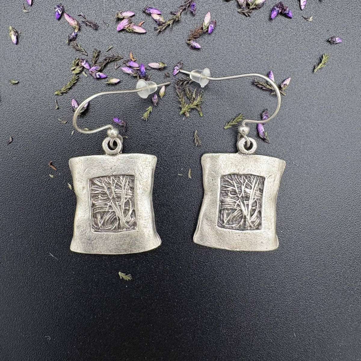 A pair of silver drop earrings with rectangular textured pendants featuring an engraved tree design, displayed on a dark background with scattered dried lavender petals.