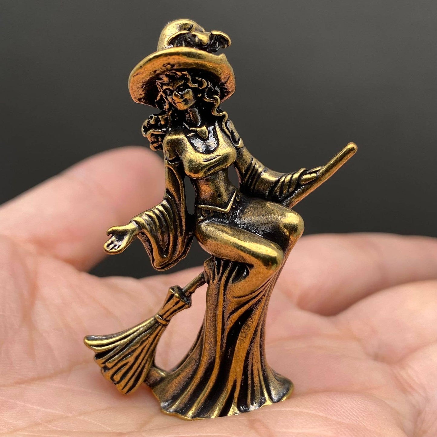 A small brass figurine of a witch seated on a broom wearing a wide-brimmed hat and flowing dress, displayed on a person's palm against a dark gray background.