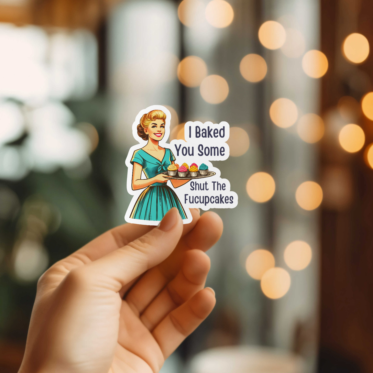 Sticker with a cartoon woman holding cupcakes and text, held by a hand against a blurred background