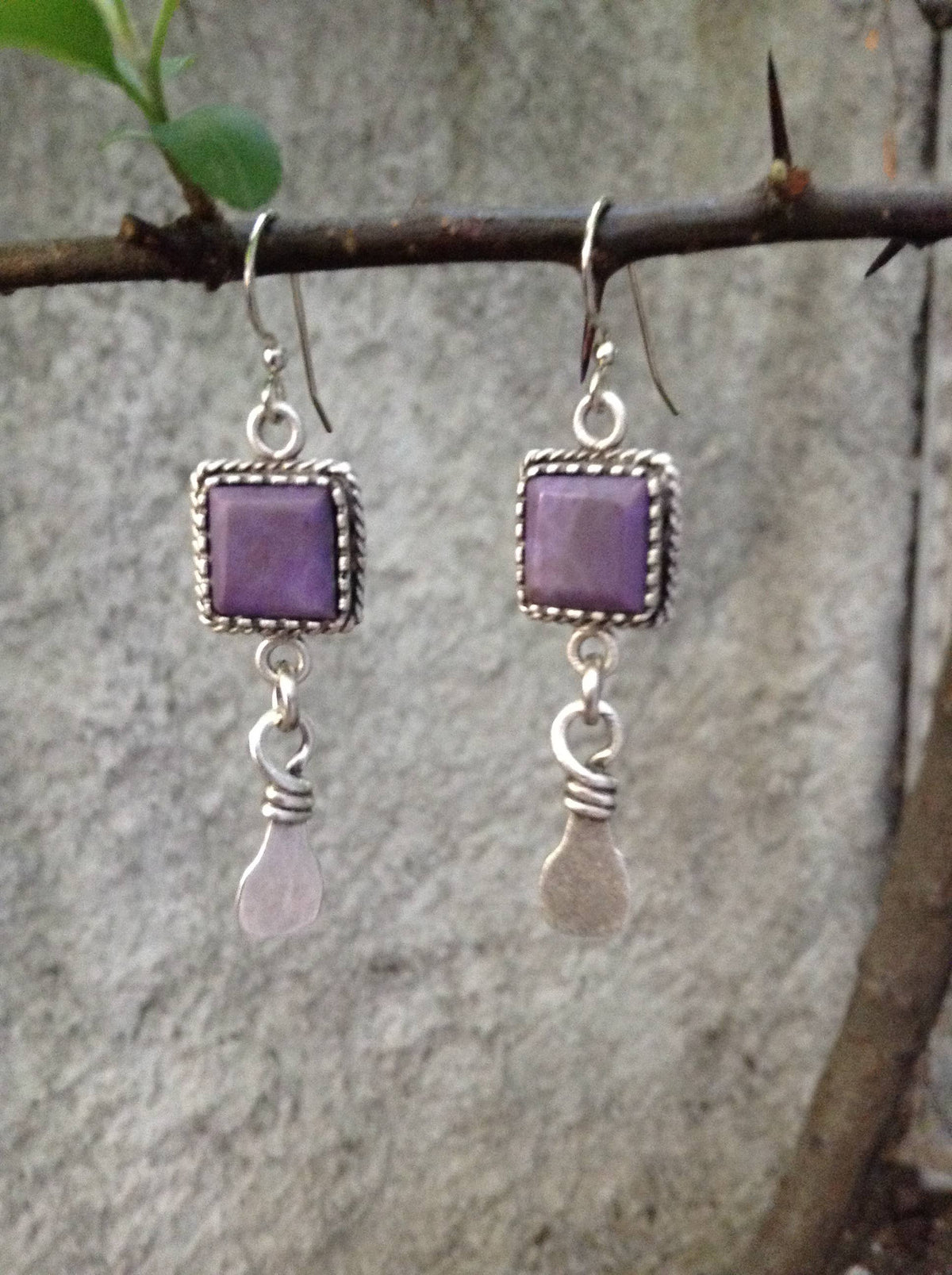 A pair of handmade dangle earrings with square purple stone tops and silver-toned wire-wrapped teardrop charms, hanging from a tree branch against a blurred gray stone wall.
