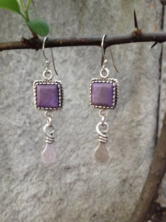 A pair of handmade dangle earrings with square purple stone tops and silver-toned wire-wrapped teardrop charms, hanging from a tree branch against a blurred gray stone wall.