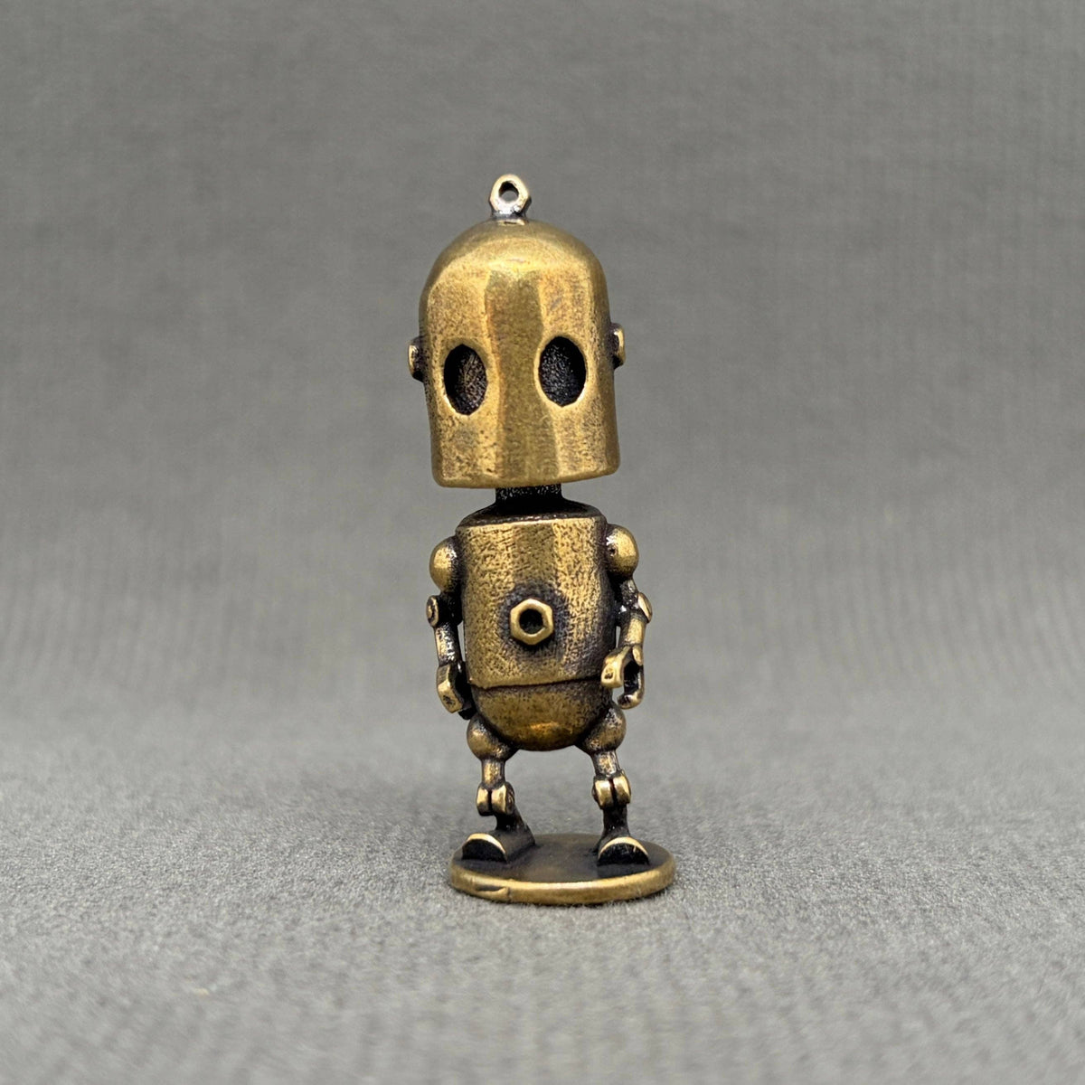 A small brass robot figurine, standing on a round base against a textured gray background.