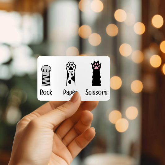 Hand holding a sticker with rock, paper, scissors cat paws against a blurred background